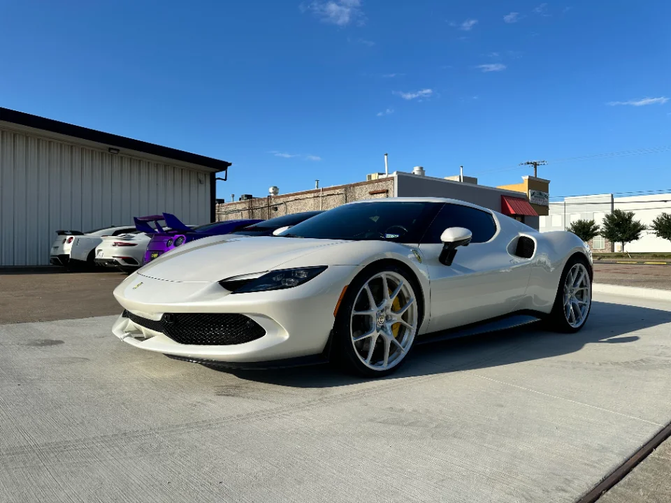 A white Ferrari 296 GTB parked outside a building with a purple Nissan GT-R and a white Porsche 911 in the background.