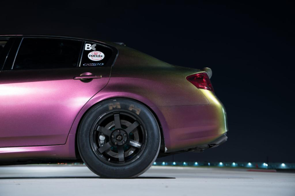 Rear quarter view of a color-shifting Infiniti G37 with drag slicks and black wheels at a drag strip at night.