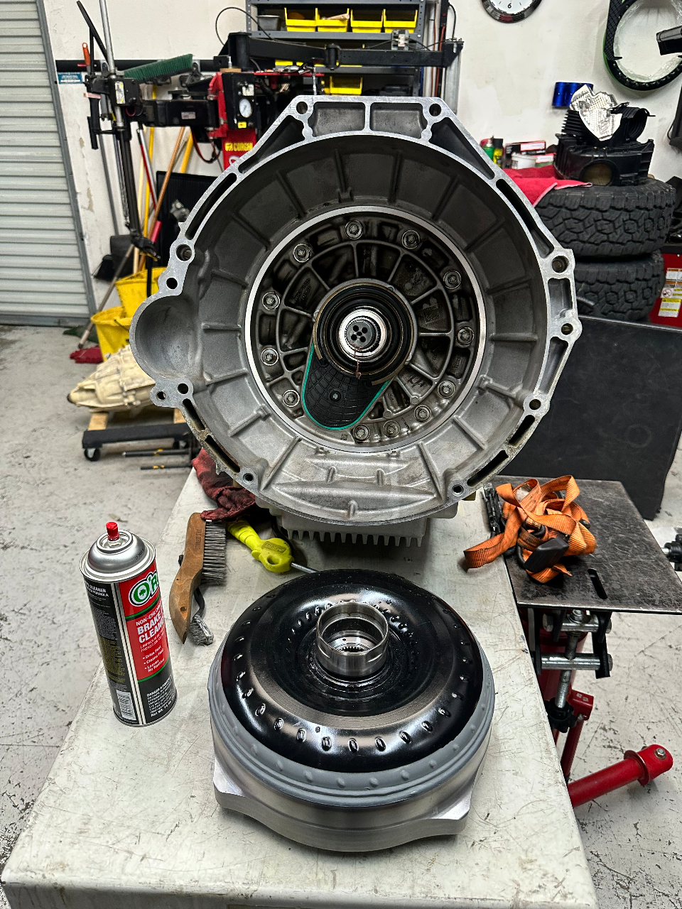 A disassembled transmission casing and torque converter on a workbench in an automotive repair shop, with tools nearby.