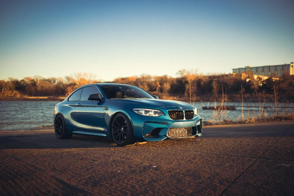 A blue BMW M2 coupe parked by a lake at sunset, showcasing its sleek design and aftermarket wheels.