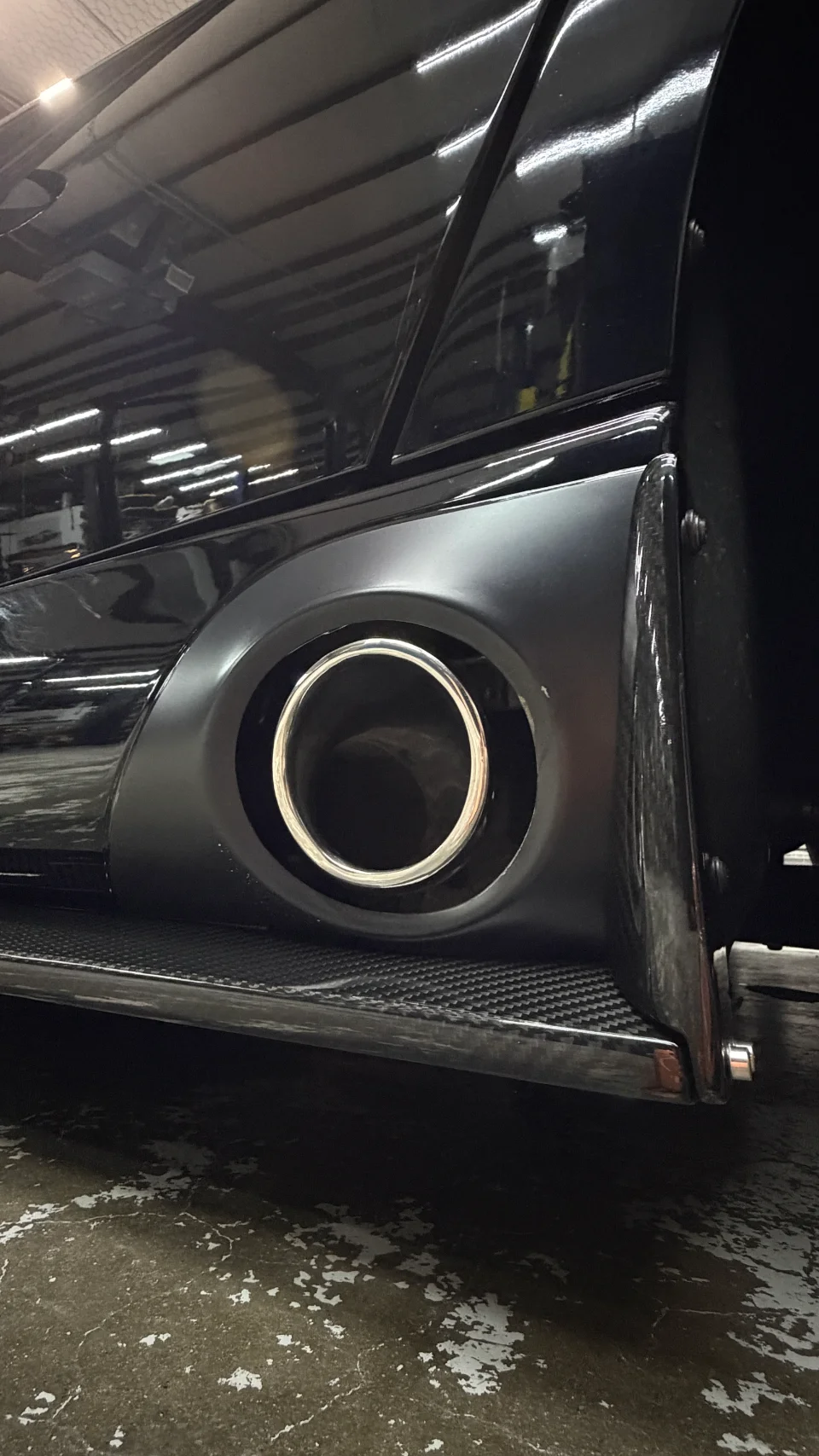 A close-up detail of a black car's custom exhaust tip and carbon fiber bodywork on a shop floor.