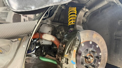A close-up view of a car's performance suspension and brake system, featuring a yellow coilover, large brake rotor, and caliper.