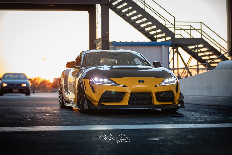 A yellow Toyota Supra with a carbon fiber hood and aero modifications on a track at sunset, with another car in the background.