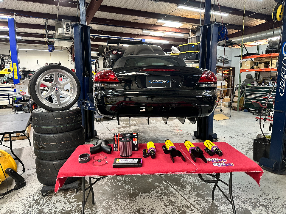 A black Mercedes-Benz CLK convertible on a lift in a Jotech Motorsports shop, with aftermarket wheels, tires, coilovers, and intake parts displayed on a table.