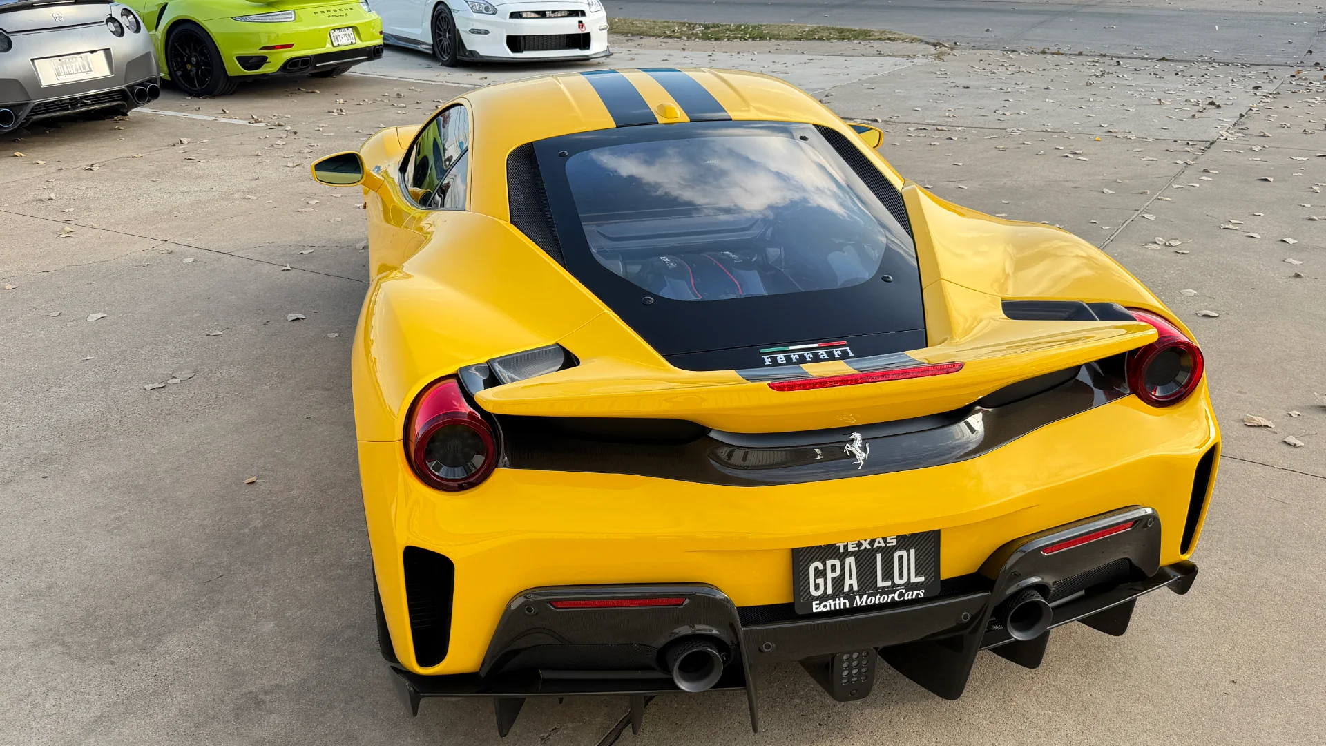 A yellow Ferrari 488 with black racing stripes and a carbon fiber diffuser, parked outdoors with other sports cars.