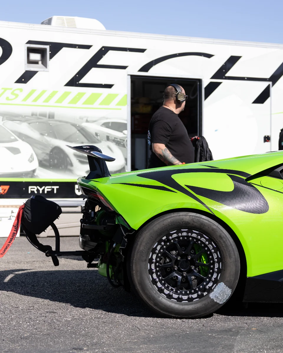 A lime green Lamborghini Huracan drag car with a parachute and drag wheels, parked next to a Jotech Motorsports trailer at a track.