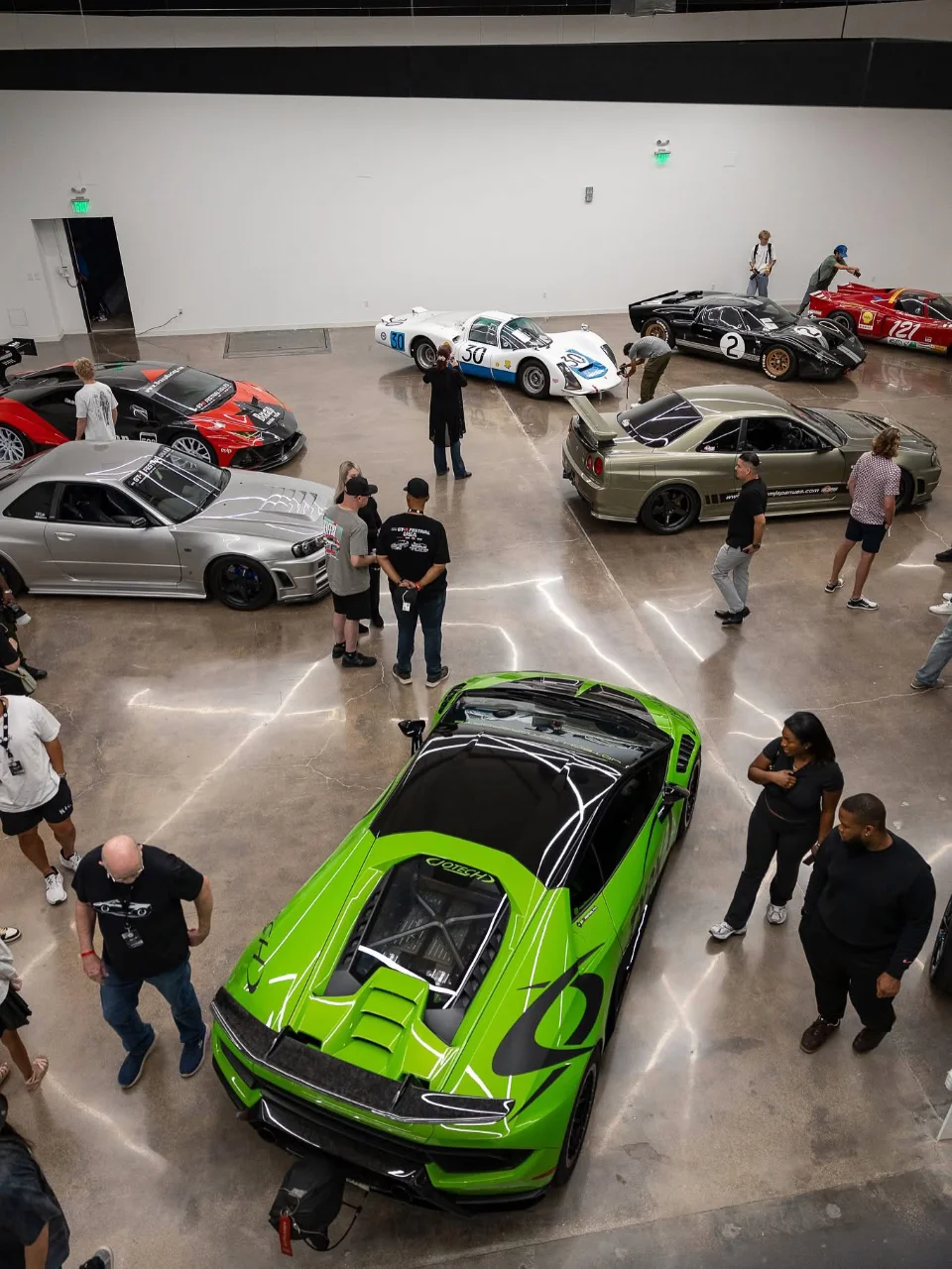 Overhead view of a car event with multiple supercars including a green Lamborghini, Nissan GT-Rs, and classic race cars, surrounded by people.