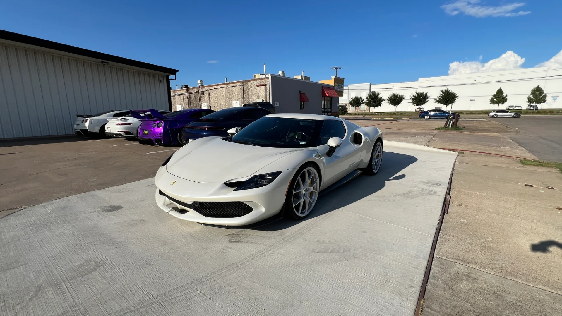 A white Ferrari 296 GTB parked in front of a building, with several Nissan GT-Rs in white and purple parked behind it.