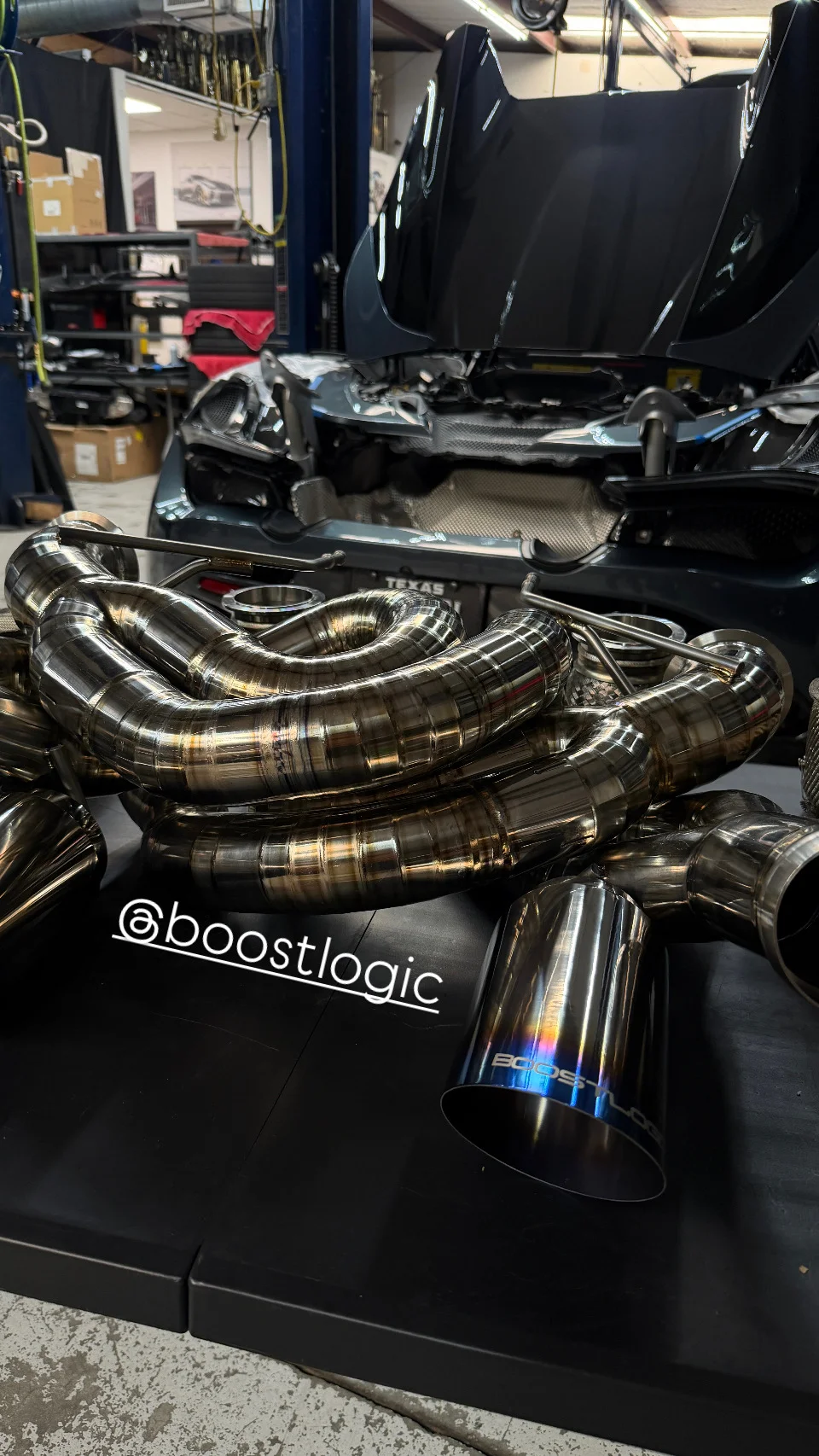 A Boost Logic titanium exhaust system with iridescent tips laid out on a table in a performance tuning shop, with a McLaren in the background.