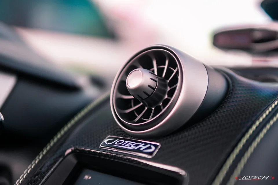 A close-up of a McLaren interior showing a metallic air vent, carbon fiber dashboard, and Jotech logo.