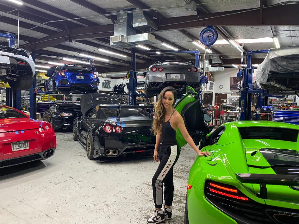 A woman stands in the Jotech Motorsports shop facility surrounded by high-performance cars including Nissan GT-Rs, a McLaren, and a Honda Civic Type R.