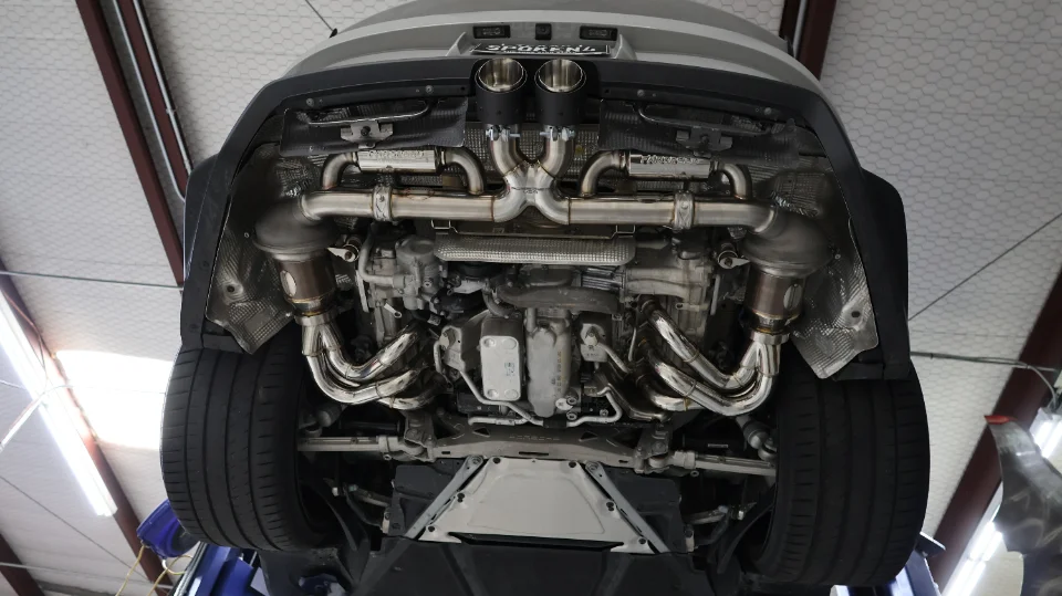 Underside view of a Porsche on a lift, showcasing a high-performance Sporrenz exhaust system with headers and catalytic converters.