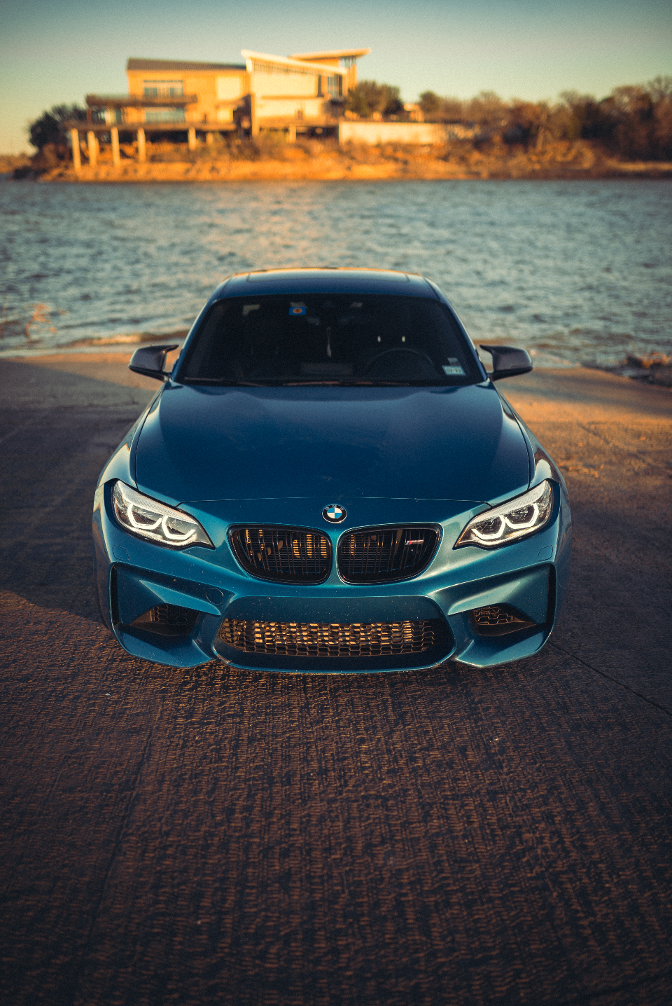 A blue BMW M2 parked on a concrete ramp next to a lake at sunset, showcasing its front end.