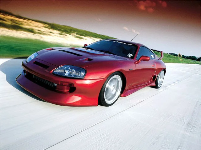 A red Toyota Supra MKIV sports car with aftermarket wheels and a large rear wing, captured in a dynamic roller shot on a road.