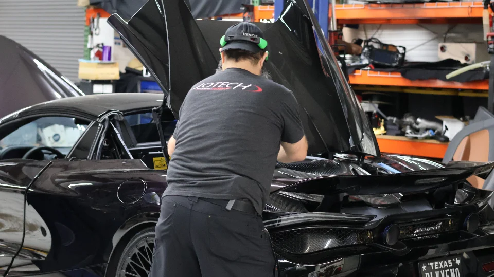 A mechanic working on the open engine bay of a black McLaren supercar in a Jotech Motorsports shop.