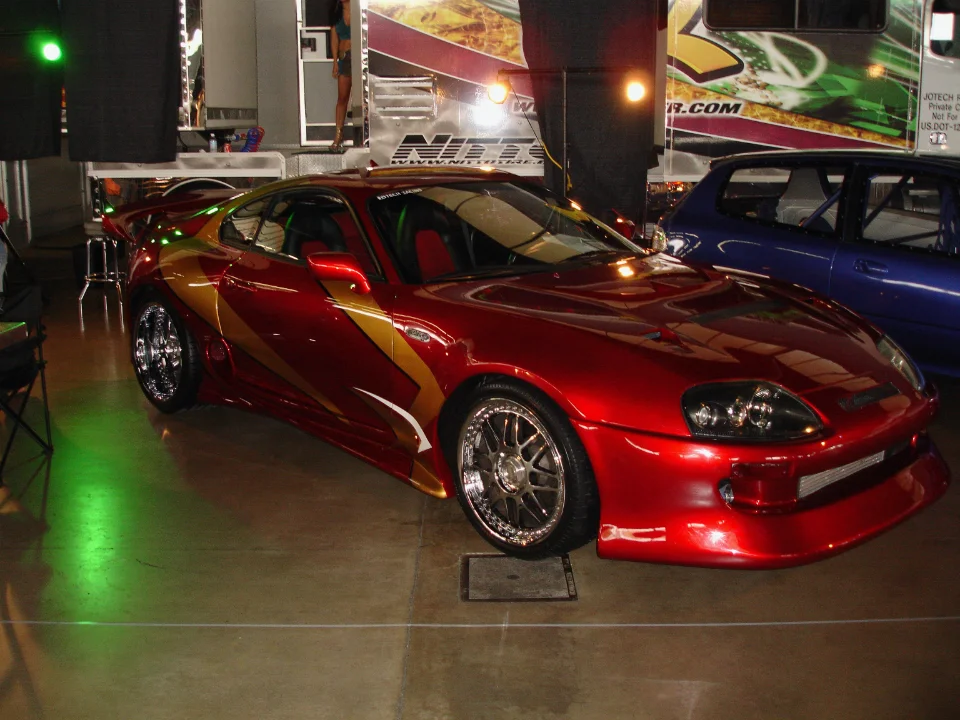 A custom red Toyota Supra with gold and white graphics and chrome wheels on display at an indoor event.