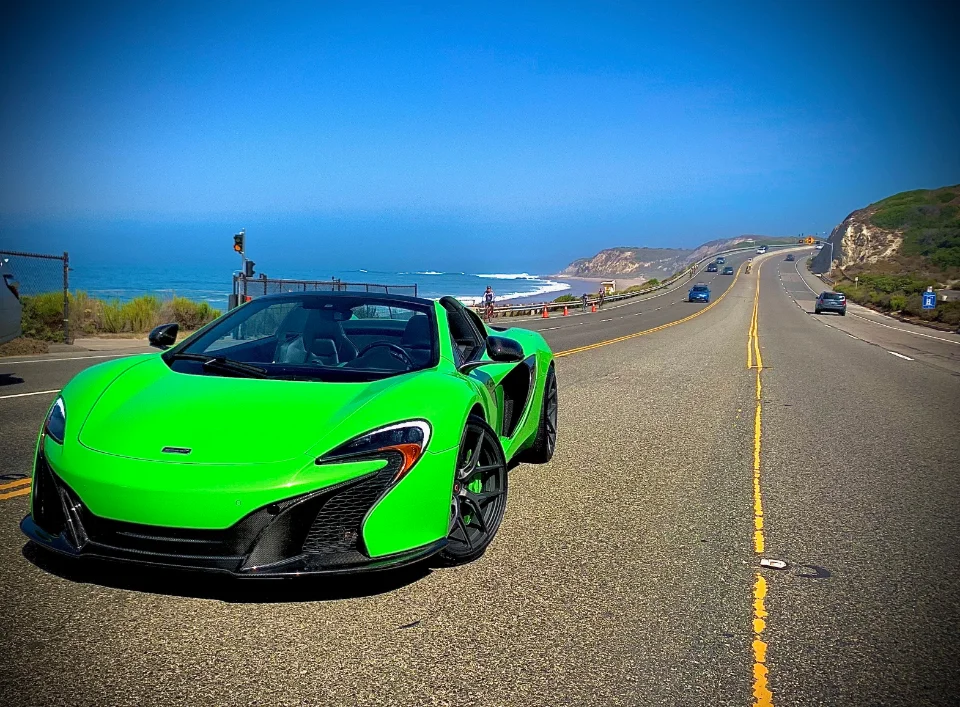 A vibrant green McLaren convertible parked on a scenic coastal highway overlooking the ocean on a clear day.