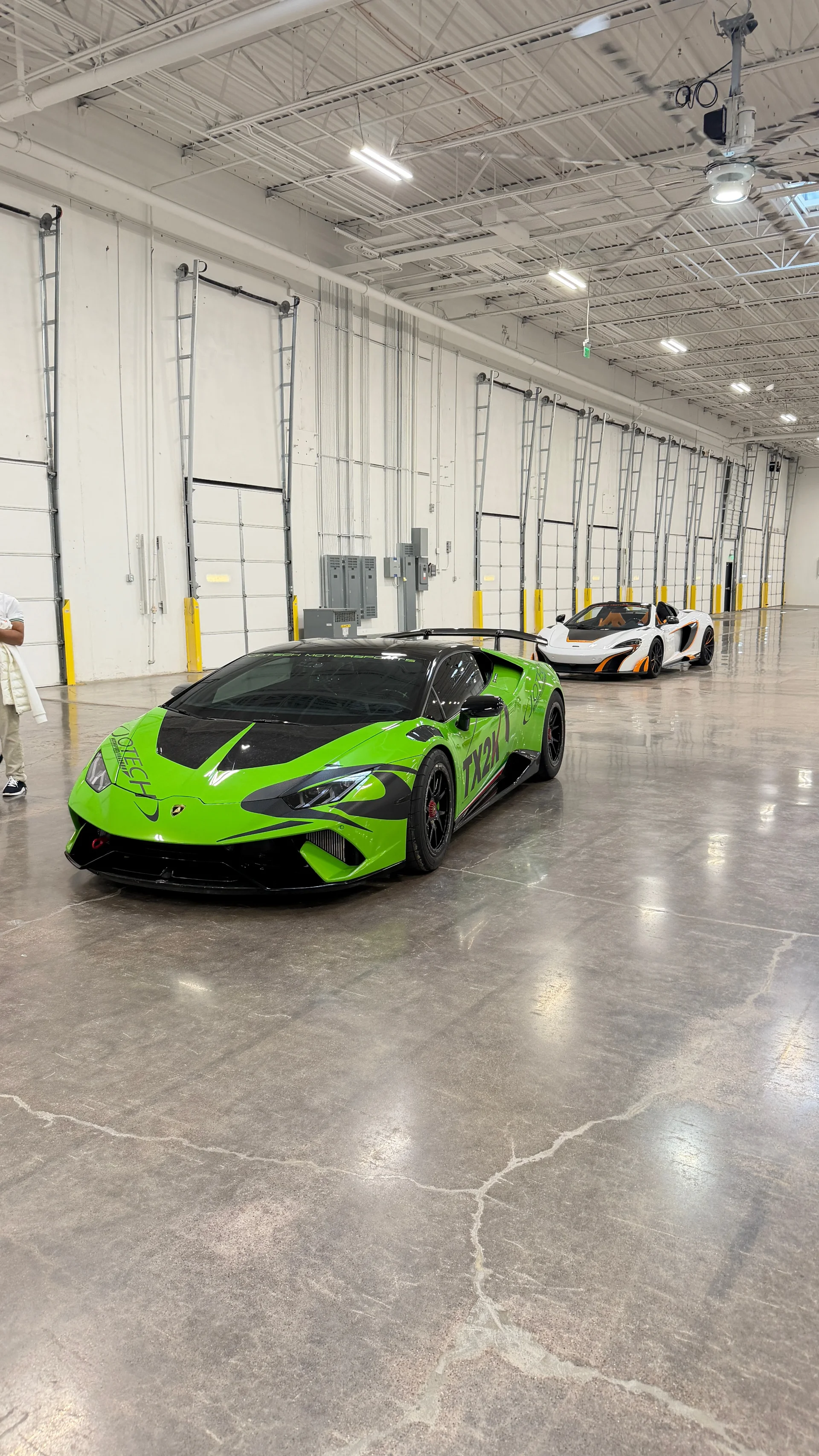 A vibrant green Lamborghini Huracan and a white McLaren supercar parked on a polished concrete floor in a large, modern facility.