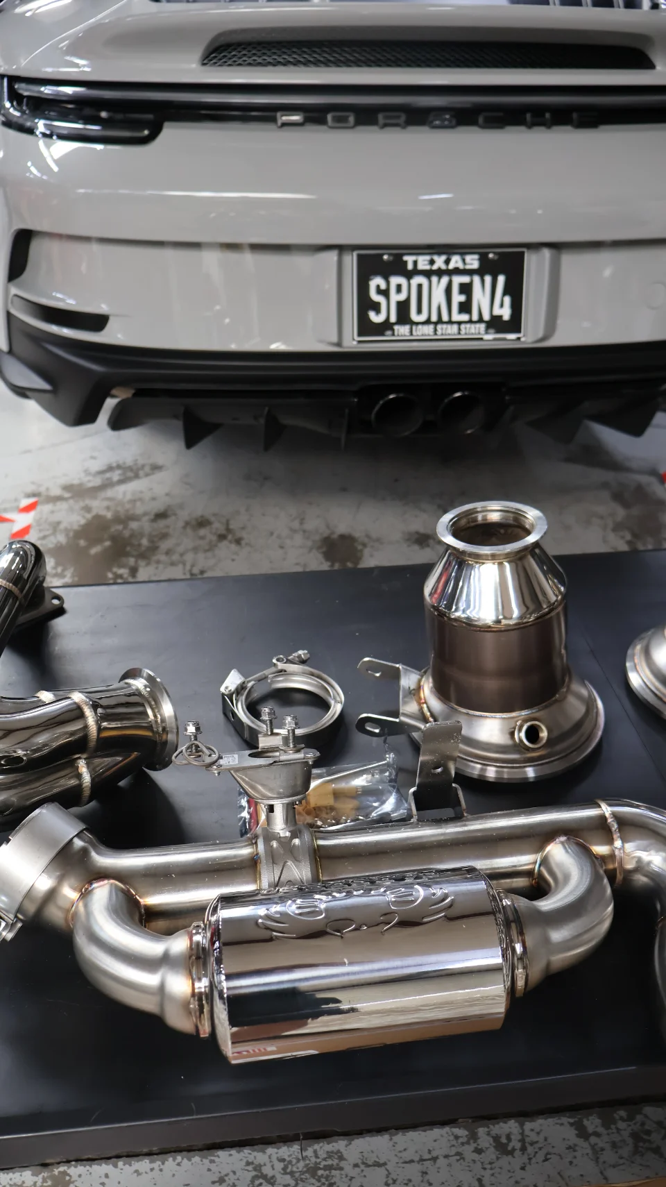 Rear view of a grey Porsche 911 with a performance exhaust system laid out on a table in a shop.