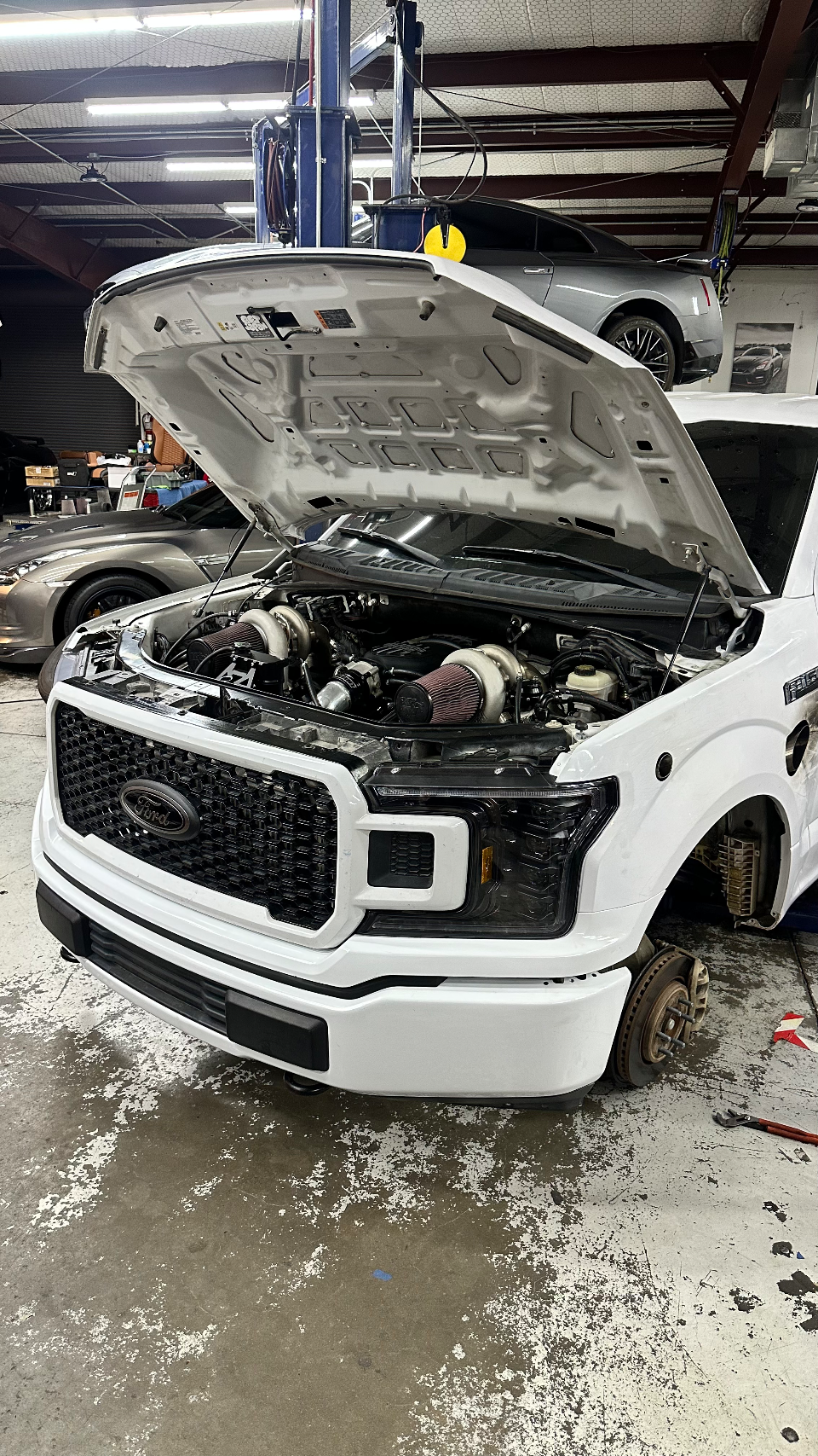 A white Ford F-150 truck with its hood open, revealing a twin-turbo engine setup, being worked on in a performance shop. A Nissan GT-R is on a lift in the backg