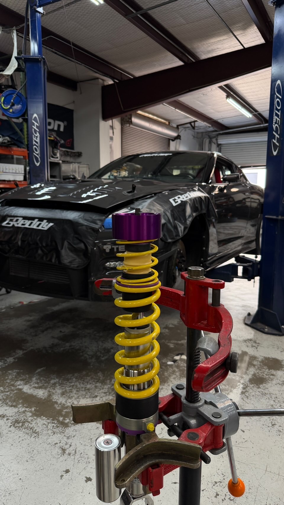 A yellow and purple coilover suspension assembly being worked on with a spring compressor in a Jotech Motorsports shop, with a Nissan GT-R on a lift in the back