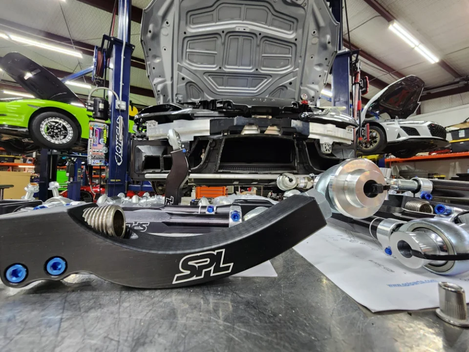 Performance suspension parts from SPL Parts on a workbench in a Jotech Motorsports shop with cars on lifts in the background.