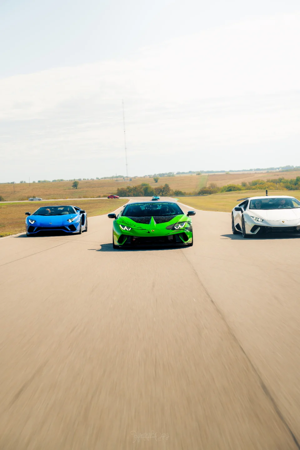 Three Lamborghini supercars, including a green Huracan and a blue Aventador, driving on a race track under a clear sky.