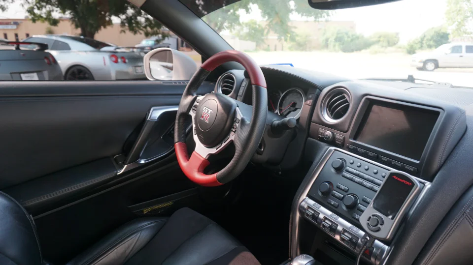 Interior view of a Nissan GT-R featuring a custom red and black steering wheel and a Cobb Accessport on the dashboard.