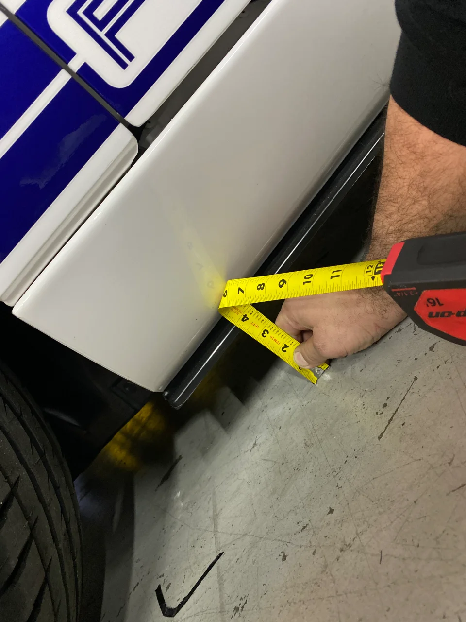 A person uses a tape measure to check the fitment of a white car's side skirt with blue racing stripes.