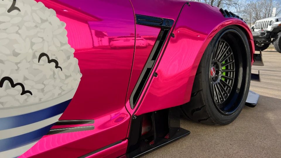 A close-up of a pink widebody Nissan GT-R with a custom rice bowl graphic, aftermarket wheels, and performance tires.