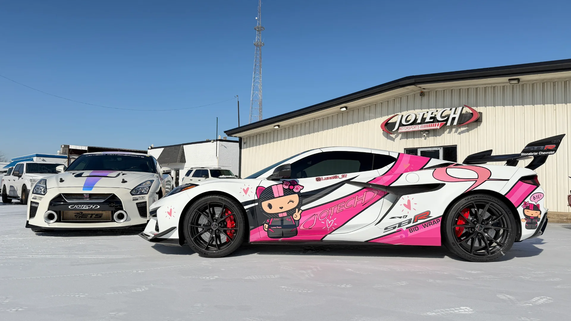 A white Nissan GT-R and a white and pink wrapped C8 Corvette parked in front of the Jotech Motorsports shop in the snow.