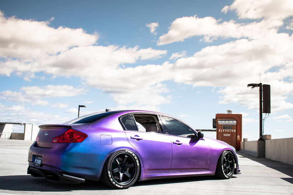 A purple color-shift Infiniti G37 sedan with aftermarket wheels parked on a rooftop parking garage under a blue sky.
