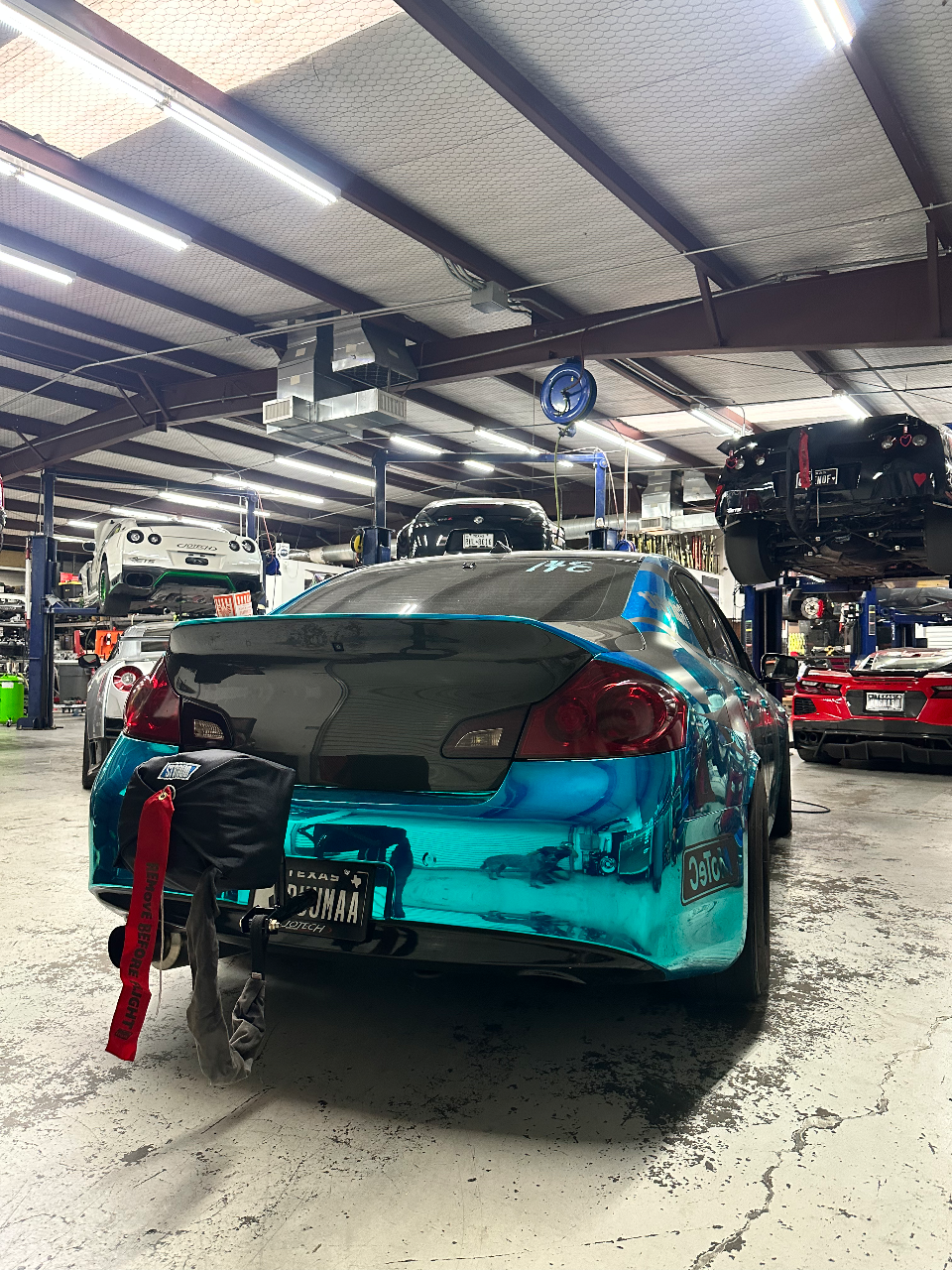 A teal wrapped Infiniti G37 coupe with a parachute mounted on the rear, parked in a performance tuning shop with other cars on lifts.