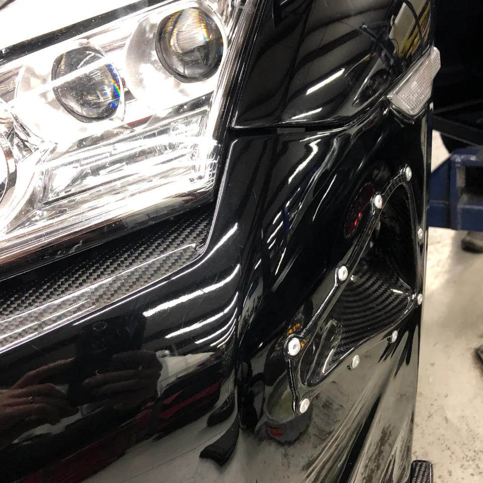 Close-up of a black car's front bumper and headlight, featuring carbon fiber trim and clear lens.