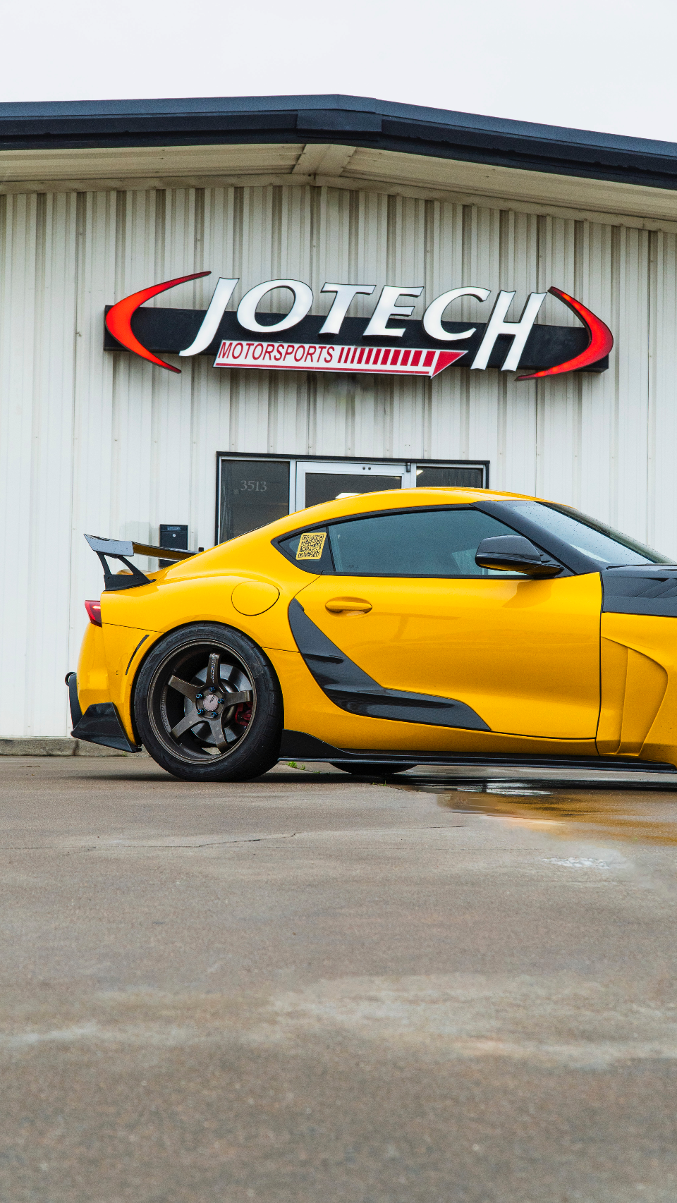 A yellow Toyota Supra with aftermarket wheels and a large rear wing parked outside Jotech Motorsports performance shop.