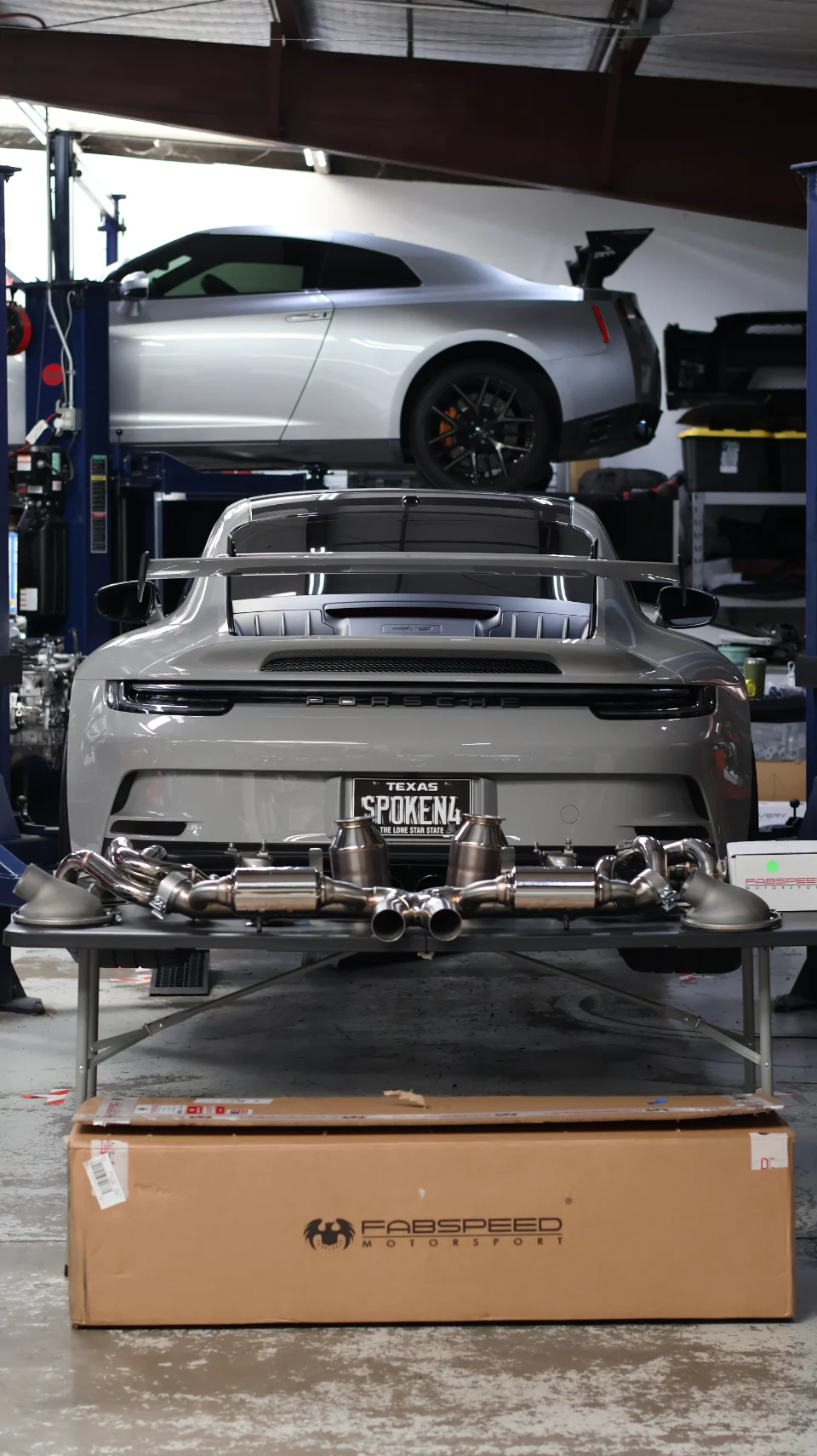 A grey Porsche 911 GT3 and a silver Nissan GT-R on lifts in a performance shop, with a Fabspeed exhaust system on a table.