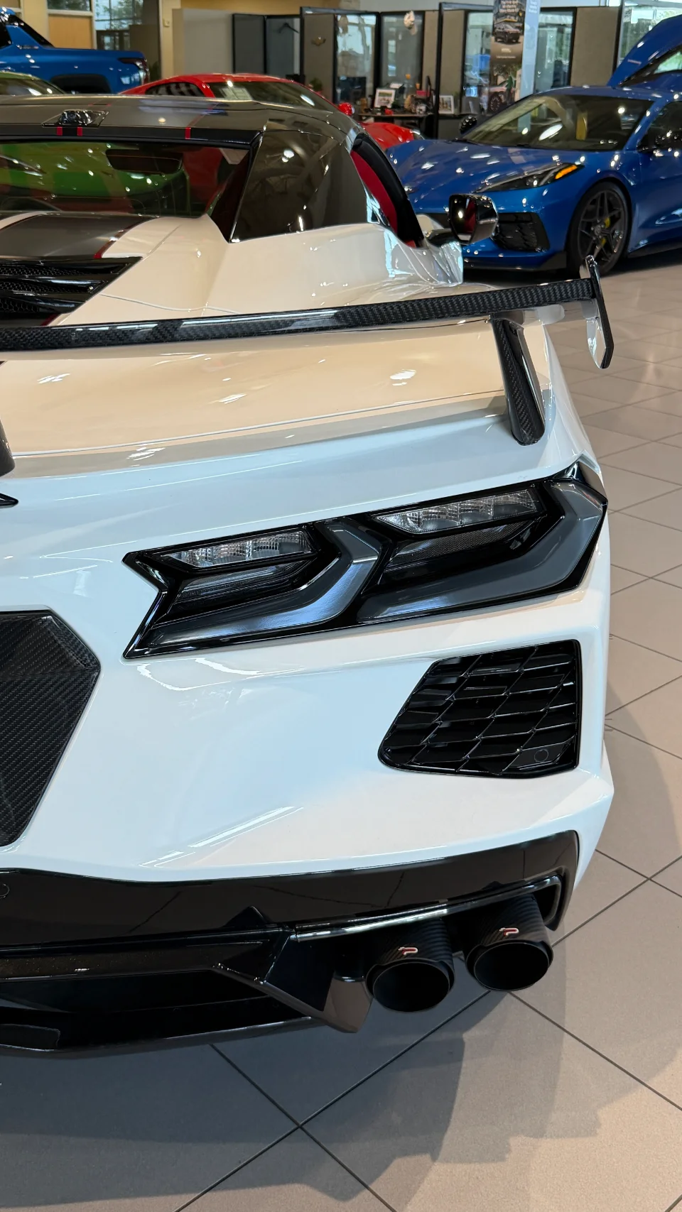 A white Chevrolet Corvette C8 Z06 with a prominent carbon fiber wing and performance exhaust tips, displayed in a car showroom.