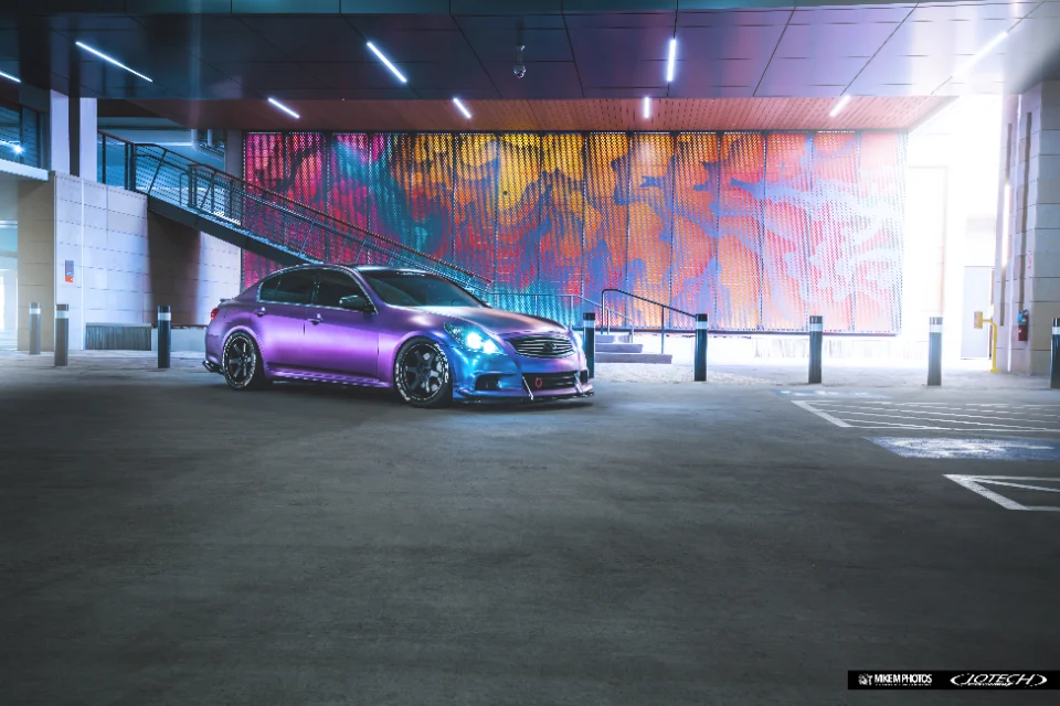 A custom purple and blue wrapped Infiniti G37 sedan with aftermarket wheels parked in an urban parking garage at night.