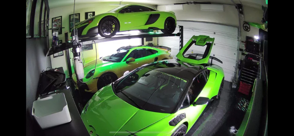 Three high-performance sports cars, including a green McLaren, a yellow Porsche, and a green Lamborghini, parked in a well-lit garage with a car lift.
