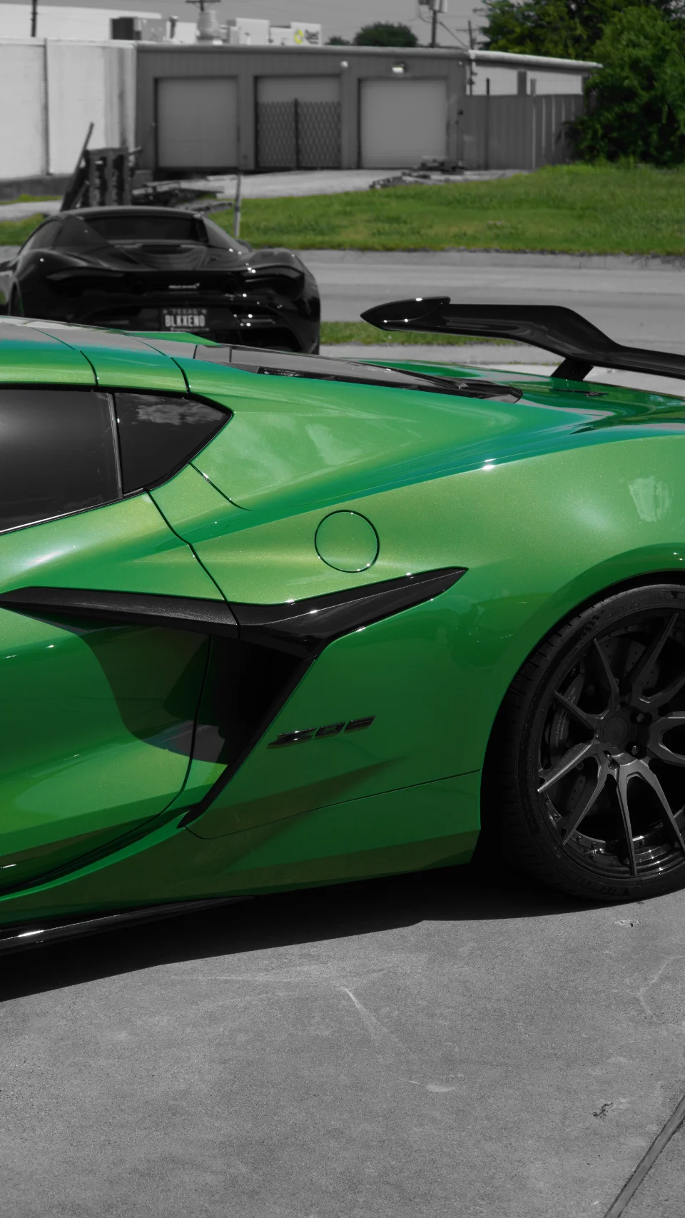 A vibrant green Chevrolet Corvette Z06 with a prominent carbon fiber rear wing and custom black wheels, parked with a black McLaren in the background.