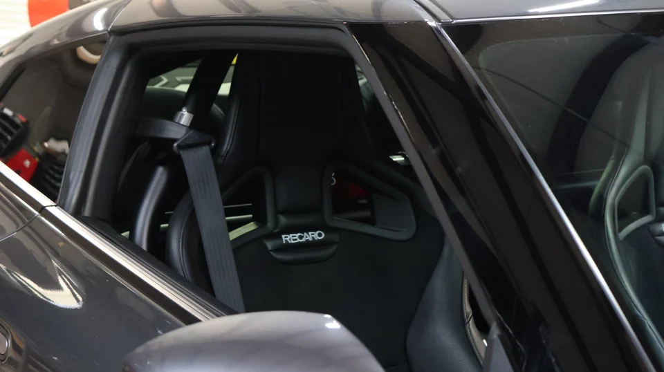 A close-up view of a black Recaro racing seat with a seatbelt and roll bar inside a performance car.