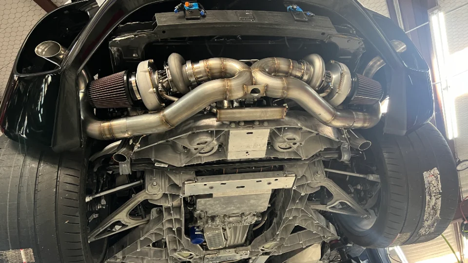 Underbody view of a black C8 Corvette showcasing a custom twin-turbo system with stainless steel exhaust piping and air filters.