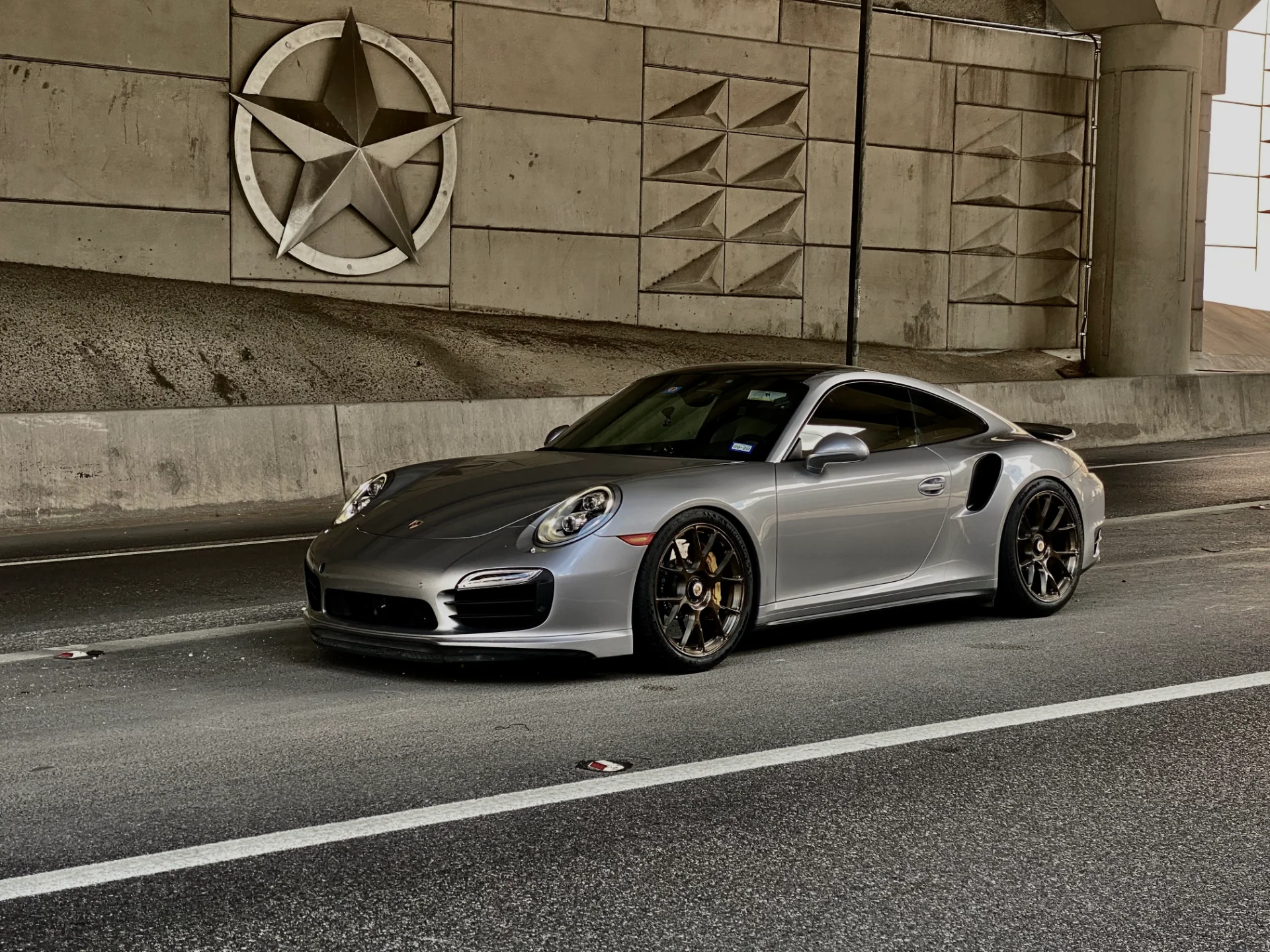 A silver Porsche 911 Turbo S with custom wheels parked under an overpass with a large Texas star emblem on the wall.