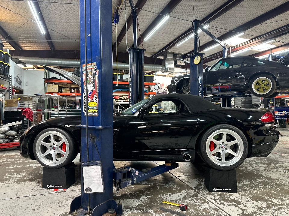 A black Dodge Viper convertible on a car lift in a performance tuning shop, with a black Porsche 911 on a lift above it.