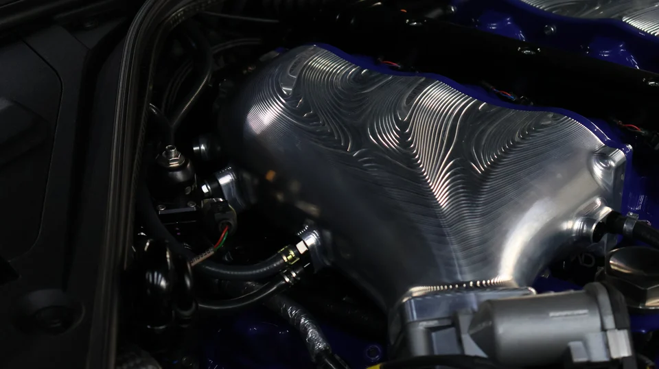 Close-up of a custom billet intake manifold with intricate machining in a high-performance car engine bay with blue accents.