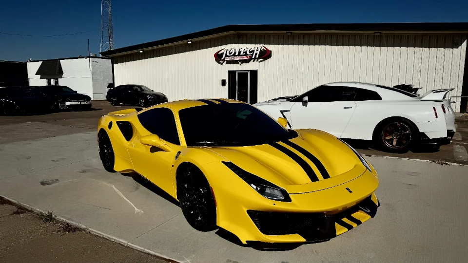 A yellow Ferrari 488 with black racing stripes and a white Nissan GT-R parked outside the Jotech Motorsports shop building.