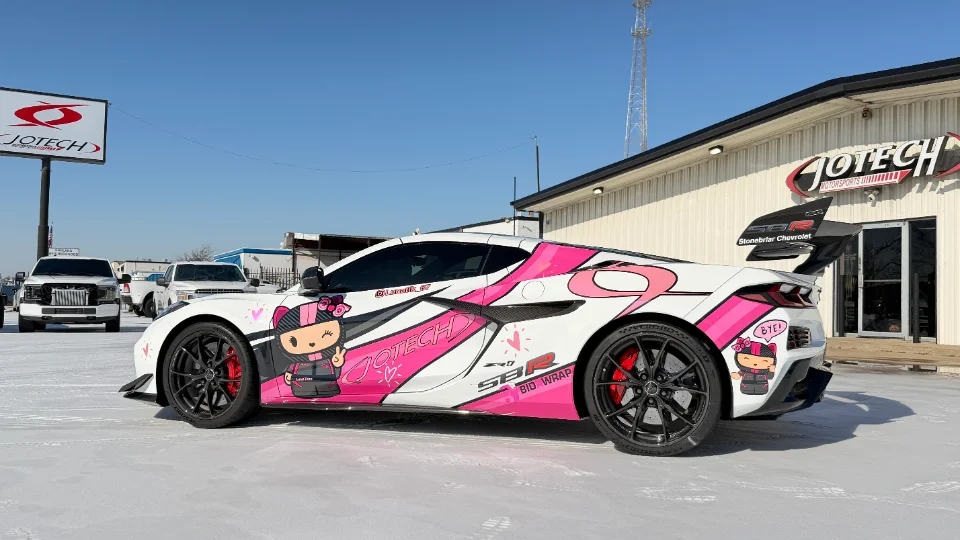 A white and pink custom-wrapped Chevrolet Corvette C8 ZR1 with a large rear wing parked outside Jotech Motorsports.