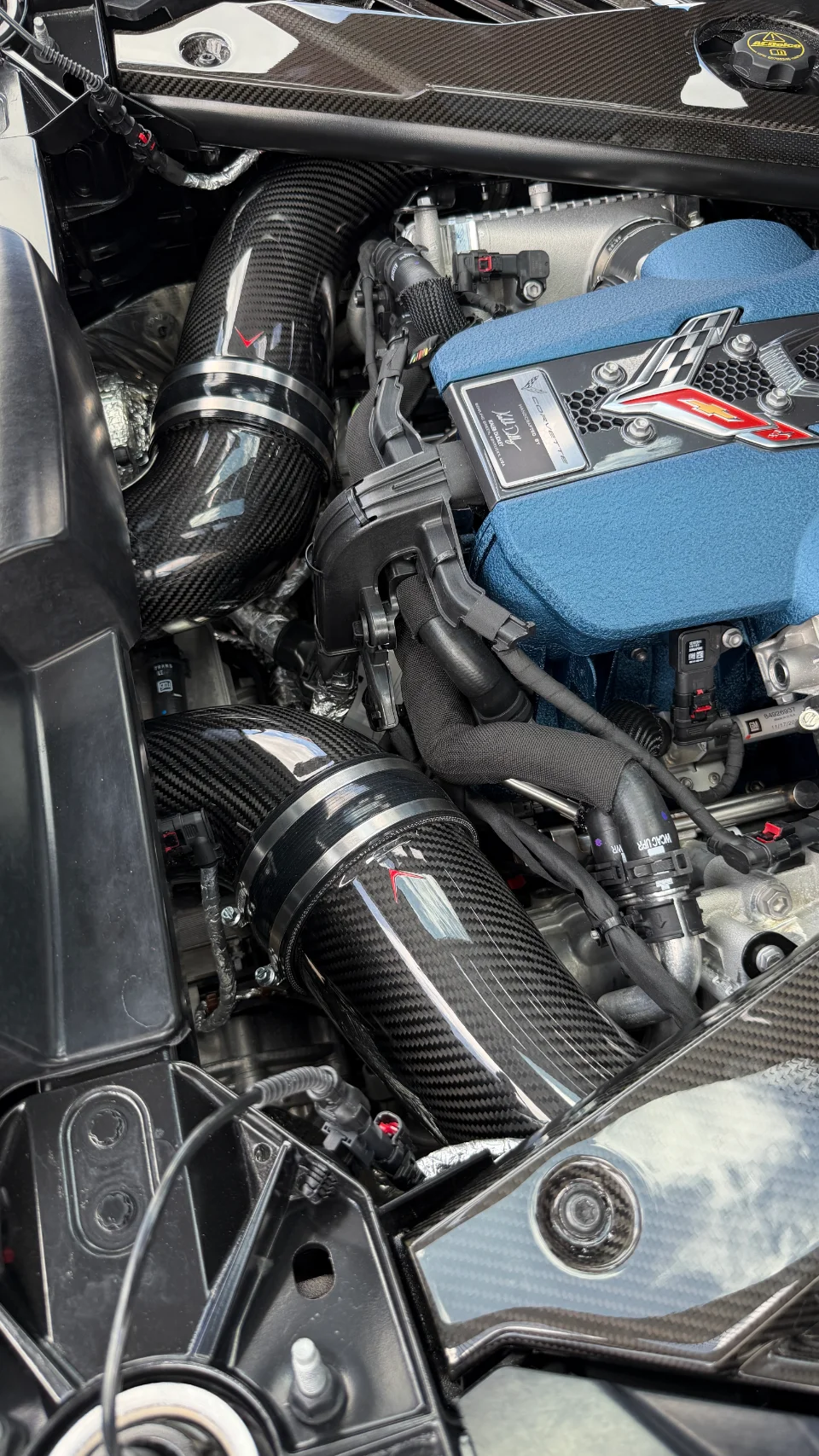 A close-up of a Corvette C8 engine bay featuring aftermarket carbon fiber intake tubes and a blue engine cover.