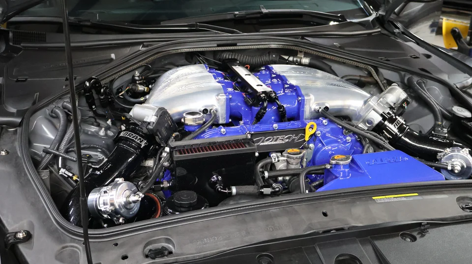 A detailed view of a modified Nissan GT-R VR38DETT engine bay featuring blue and silver aftermarket induction and boost components.