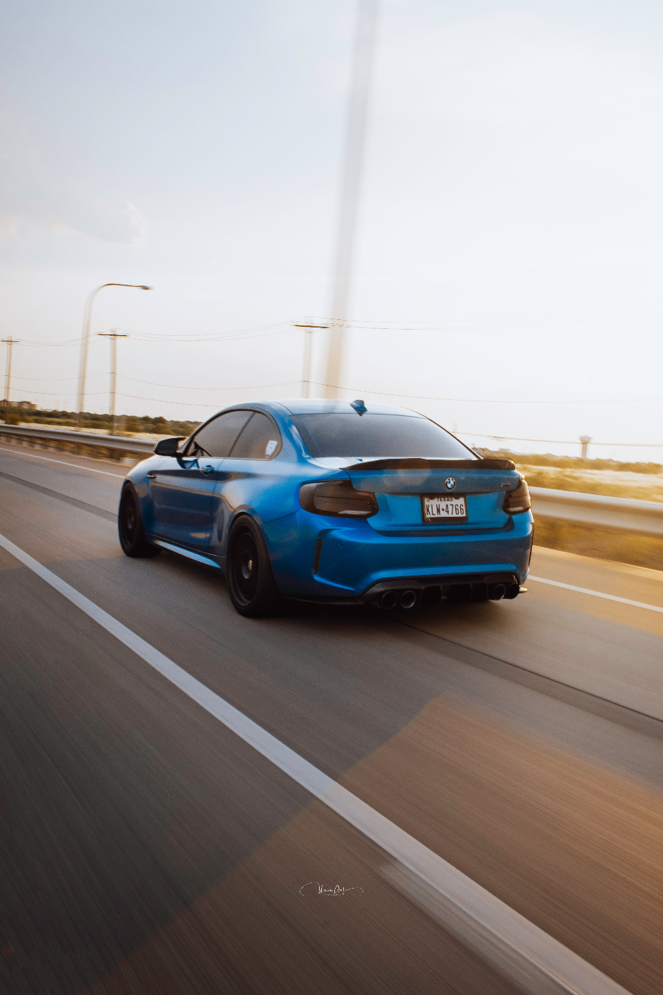 Blue BMW M2 coupe with a carbon fiber spoiler and aftermarket quad exhaust driving on a highway at sunset.